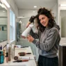 Woman styling her hair in bright modern bathroom with vanity lights and products visible