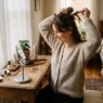 Woman styling her hair at bedroom dressing table with morning light streaming through curtains
