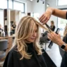 Professional stylist cutting blonde layered hair in modern salon with bright lighting and cutting cape