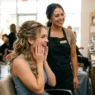 Woman admiring her completed bridesmaid half up half down hairstyle in salon mirror with stylist behind her