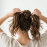 Woman demonstrating messy bun creation process with hands visible styling brown hair against white background