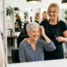 Woman over 60 with silver pixie cut smiling at her reflection while stylist stands behind her in bright salon