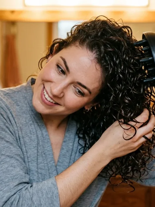 Woman using diffuser attachment to dry wavy hair with head tilted sideways in bathroom