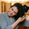 Woman using diffuser attachment to dry wavy hair with head tilted sideways in bathroom