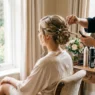 Bride in silk robe sitting while professional hair stylist works on updo styling during early morning wedding preparation