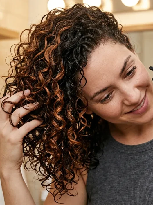 Woman scrunching curly hair with diffuser attachment, head tilted sideways in warm bathroom lighting