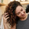 Woman scrunching curly hair with diffuser attachment, head tilted sideways in warm bathroom lighting
