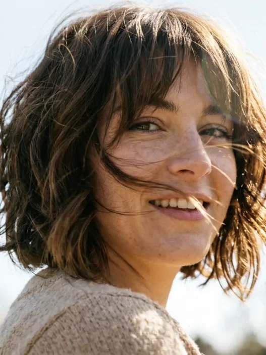 Woman with chin-length shaggy French bob and curtain bangs with hair moving in breeze under bright sky